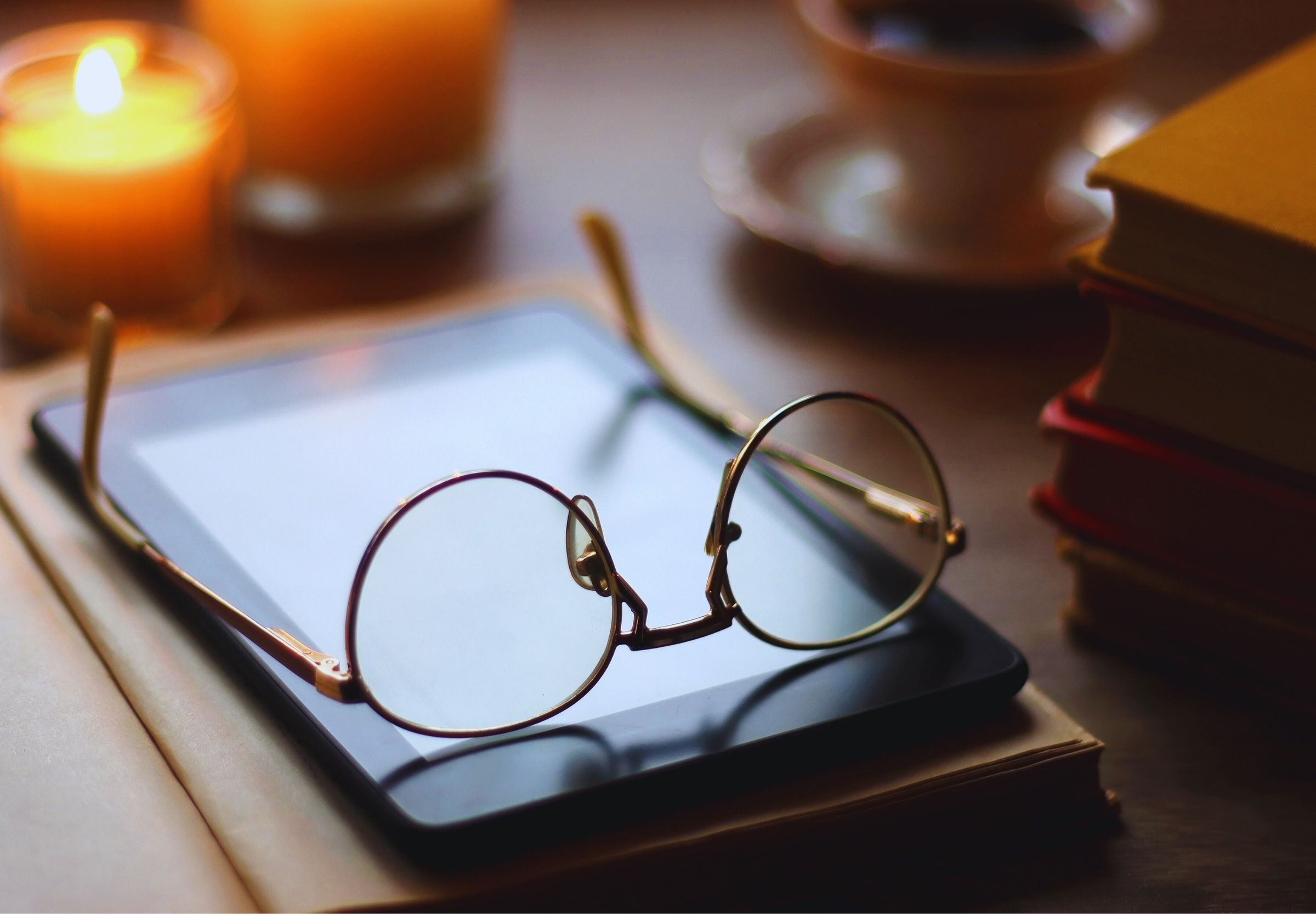 Okulary spoczywające na ekranie czytnika e-booków, w otoczeniu świec i filiżanki kawy, symbolizujące komfort i relaks podczas czytania cyfrowych książek.